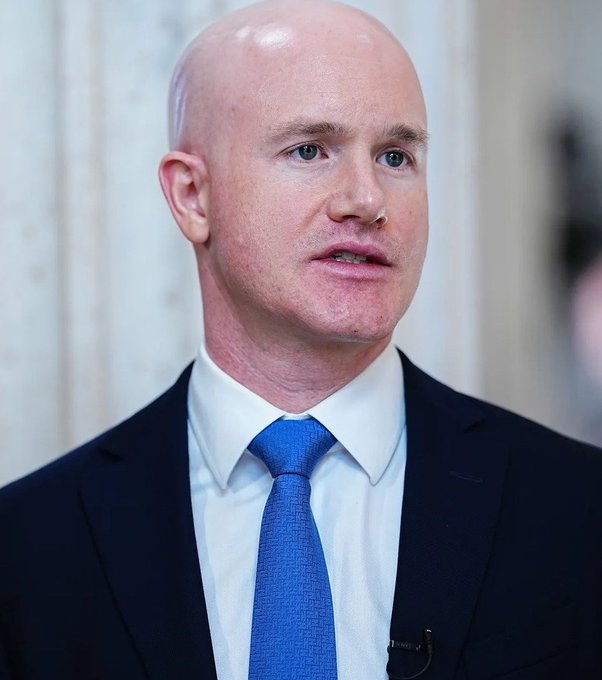 A close-up portrait of a bald man in a black suit, white shirt, and blue tie, seemingly speaking at an event.