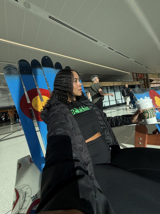 A selfie-style photo of a woman with curly hair in a black puffer jacket holding a Starbucks coffee cup while sitting in a chair decorated with the Colorado flag design.