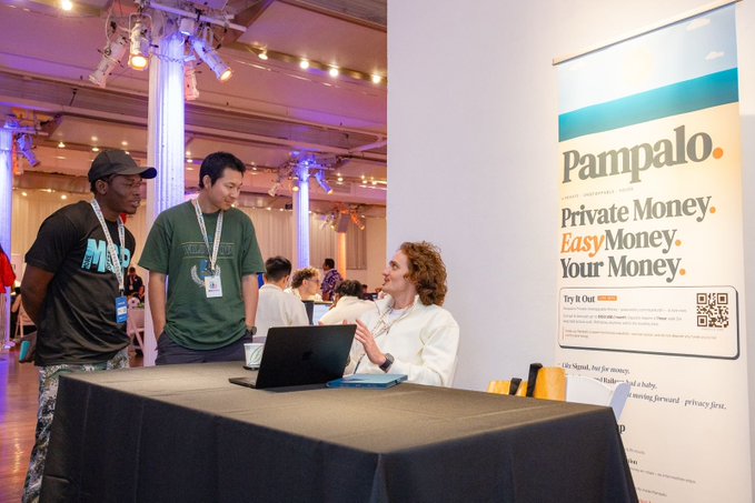 The image shows a group of people gathered around a table, with one person sitting at the table using a laptop while others stand nearby engaged in conversation. The scene appears to be set up for an event or presentation related to private money or cryptocurrency. A banner is displayed on the wall behind them, which reads "ETHGlobal New York Spotlight: @pampalodotcom Privacy-first banking that keeps your encryption keys on your own device!".