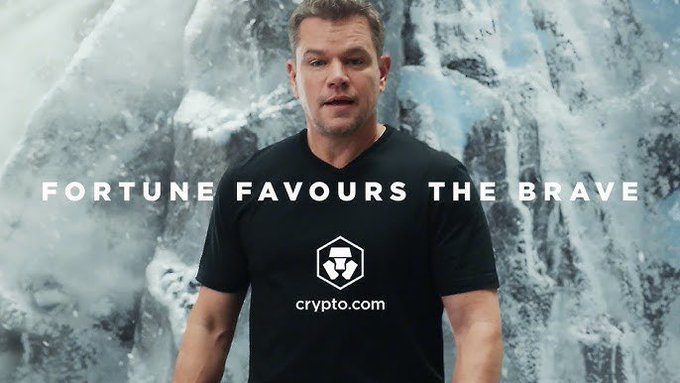 The image shows actor Matt Damon wearing a black Crypto.com t-shirt with the tagline "FORTUNE FAVOURS THE BRAVE" against an icy mountain backdrop, serving as a promotional advertisement for the cryptocurrency platform. The post humorously contrasts outrage over casting choices with the absurdity of Damon—famous for playing Odysseus in *The Odyssey*—promoting a crypto company.