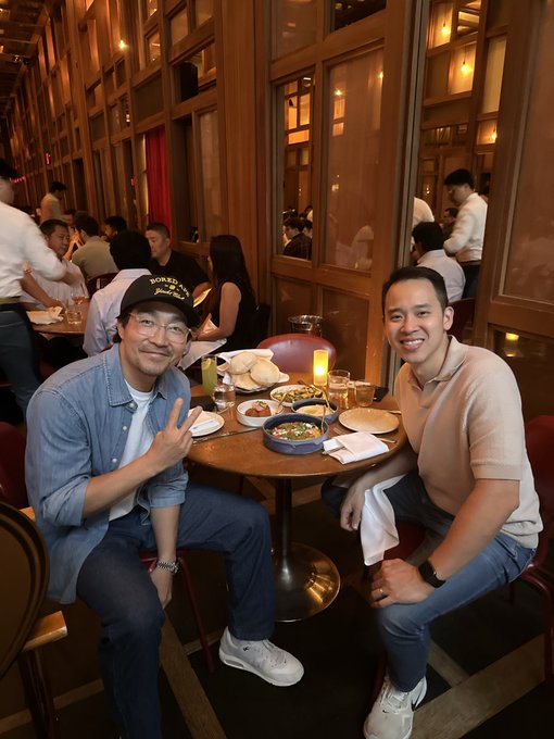 Two smiling men pose at a restaurant table filled with dishes and drinks; the man on the left wears a denim shirt and 'MURRAY'S' cap while making a peace sign.