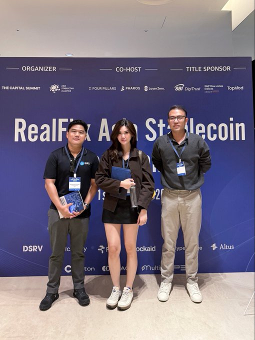Three individuals posing at a "RealFi x Asia Stablecoin" event, standing in front of a sponsor backdrop featuring logos for The Capital Summit, Asia Blockchain Alliance, Pharos, Layer Zero, SBI DigTrust, TopMed, DSRV, RockAId, Hyperlane, and Altus.