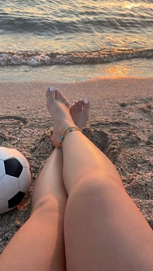 A person's bare legs and feet with white toenail polish resting on sandy beach next to a soccer ball, with ocean waves visible.