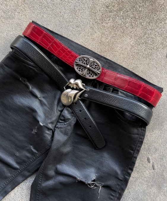 Two belts, one red crocodile-textured with an ornate silver buckle and one black leather with a gold buckle, are layered over distressed black pants on a concrete surface.