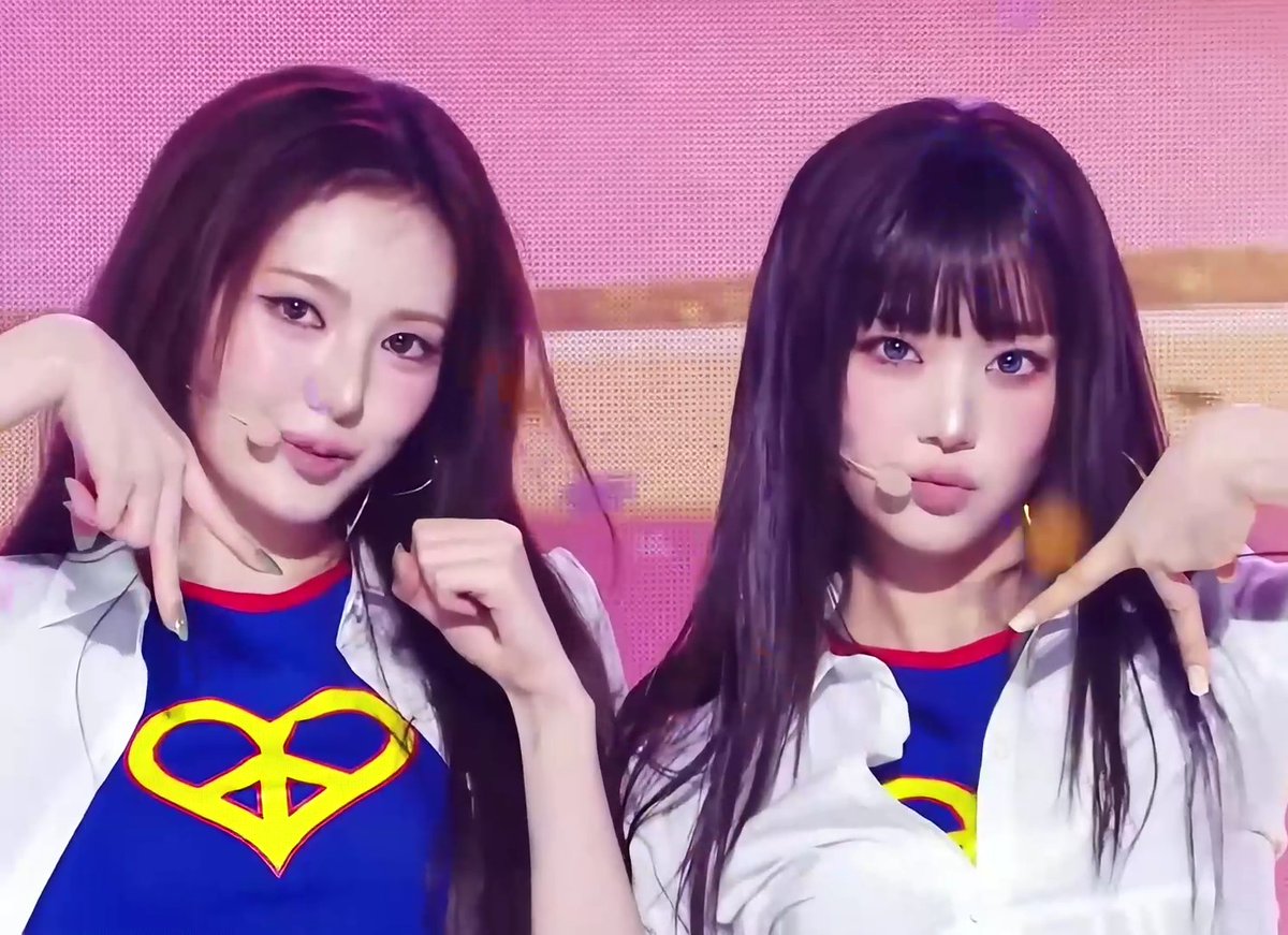 Two young women with long dark hair pose together wearing matching Superman logo t-shirts and white jackets, with pink confetti or hearts in the background.