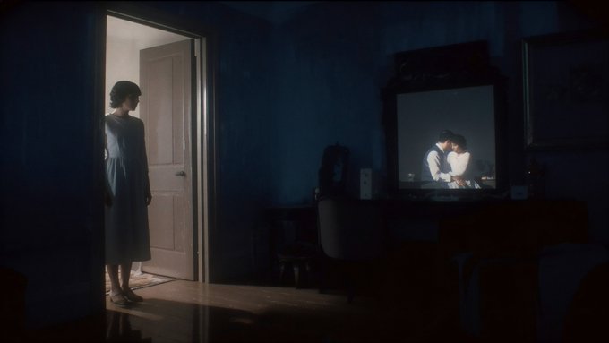 A woman in a dress stands in a doorway of a dimly-lit room, observing a couple kissing on a television screen.