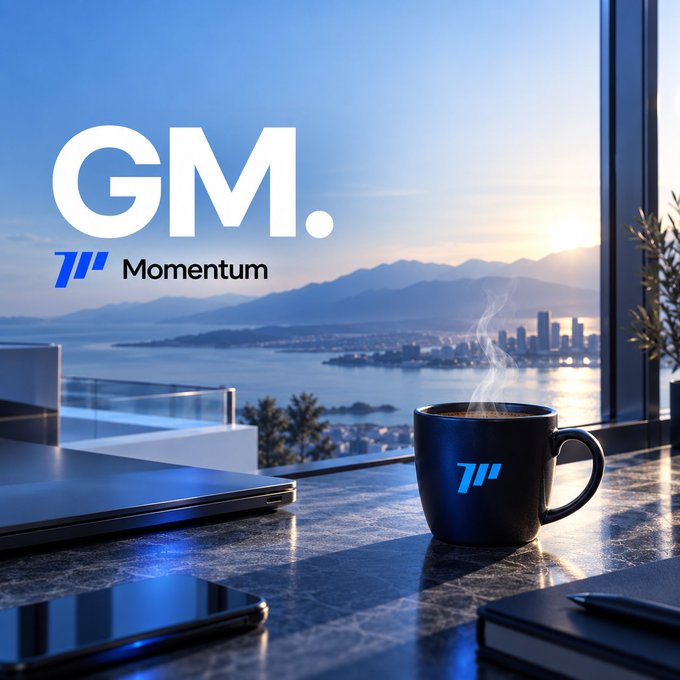 A steaming coffee mug bearing the "M" logo sits on a dark marble desk with a coastal city skyline and sunset visible through floor-to-ceiling windows, perfectly matching the relaxed, motivational morning vibe of the post. The bold "GM." text and "Momentum" branding overlay tie the scenic work setting to the upbeat Friday energy referenced in the caption.