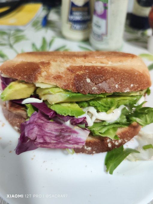 The image shows a sandwich with lettuce, avocado, and purple cabbage sitting on top of a white plate. The sandwich appears to be made with whole wheat bread, giving it a hearty appearance. In the background, there are two bottles of mayonnaise placed next to each other, suggesting that they might be used as condiments for the sandwich.