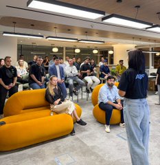 A modern office or co-working space with people gathered around yellow bean bags and seating, while a woman in a dark blue t-shirt and light jeans stands addressing them.