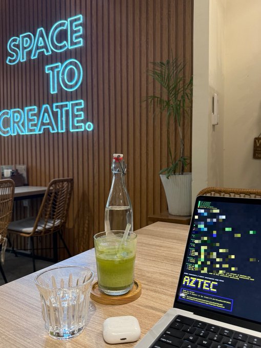 A casual café setting with a neon "SPACE TO CREATE" sign on a wood-paneled wall; a table holds a glass of green juice, a water bottle, AirPods case, and a laptop displaying code for "AZTEC".