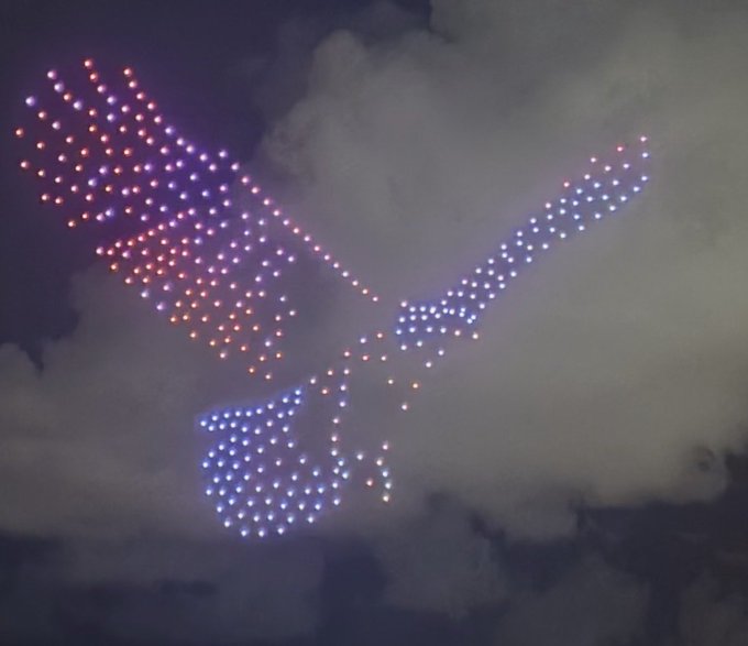 A nighttime drone light show forms the shape of a flying bird with outstretched wings, its body glowing in blue-white LEDs and one wing tinted in warm pink and orange hues against a cloudy sky. The "Gm" (good morning) post pairs with the dawn-like spectacle of the illuminated bird soaring through the morning clouds.