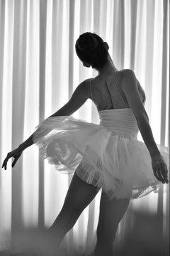 A graceful black-and-white photograph of a ballerina in a tutu posing in front of flowing sheer curtains.