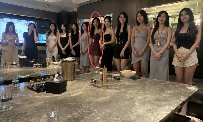 A photograph of approximately ten young women in dresses standing behind a marble countertop, with one woman in a red dress highlighted by a red oval circle.