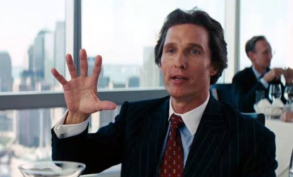 A man in a pinstripe suit and red tie gestures with his hand raised in what appears to be a restaurant setting with city views.