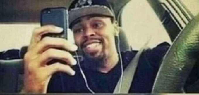 A grainy photo of a smiling young man wearing a cap and earphones, taking a selfie with a smartphone inside a car.