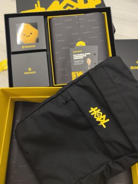 A smiley-faced yellow Binance character and matching notebook sit inside an open gift box alongside a black drawstring pouch embroidered with "9X," featured in a post celebrating Binance's 9th anniversary. The "9X" pouch aligns with the commemorative anniversary theme, as noted in the caption $BNB.