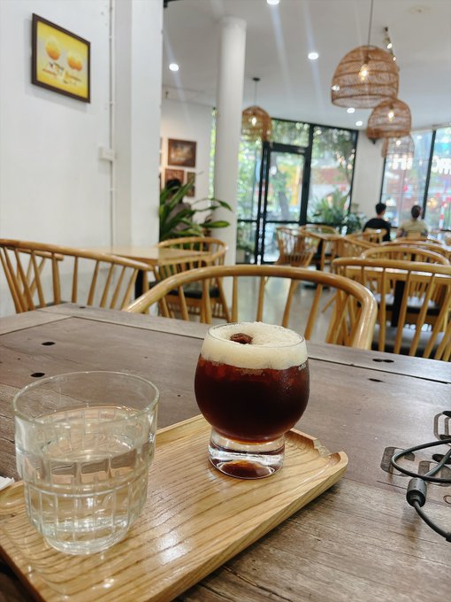 A close-up of a wooden table in a bright cafe featuring a glass of water and a coffee drink topped with foam served on a wooden tray, with wicker chairs and large windows visible in the background.