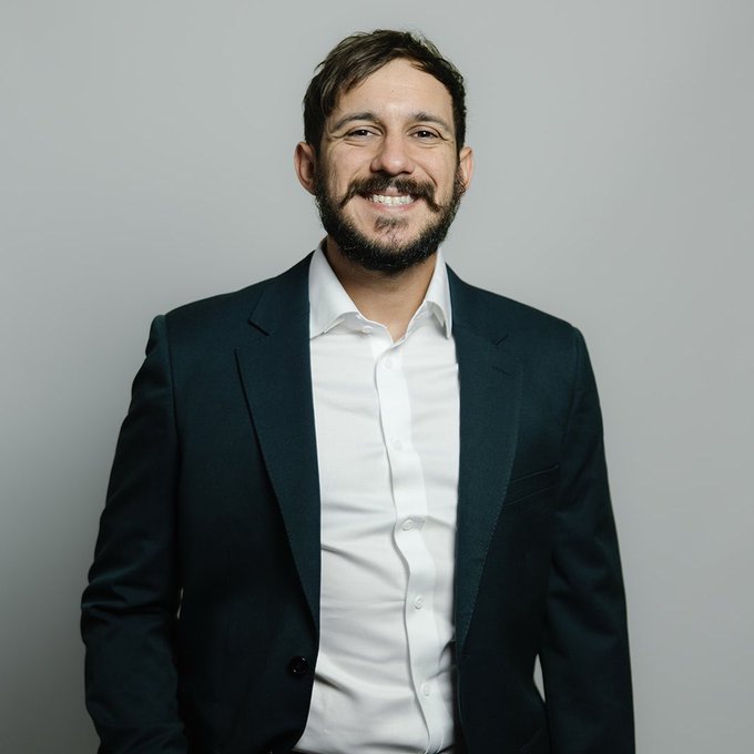 A smiling bearded man in a black blazer and white shirt poses for a professional portrait against a gray background.