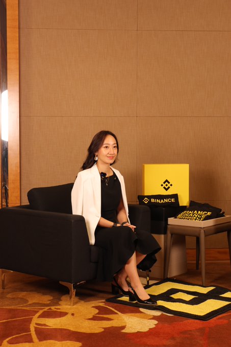 A woman sits in a black armchair at the Binance booth during TOKEN2049, with the company's logo prominently displayed on branded merchandise nearby. The scene captures the interview setting referenced in the post, where she discusses Binance's commitment to delivering lasting value beyond just generating attention in the crypto space.