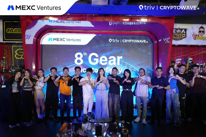 A group photo of people posing in front of an illuminated MEXC Ventures stage with '8th Gear' and 'triv | CRYPTOWAVE' branding visible in the background.