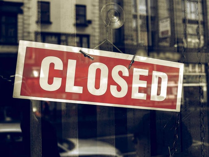 A red 'CLOSED' sign hanging in the window of a storefront, with the reflection of a city street visible in the glass.