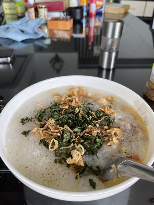 A white bowl filled with a white congee or porridge dish topped with fried shallots and dark green herbs, with a spoon resting in it, placed on a black reflective table with various kitchen items in the background.