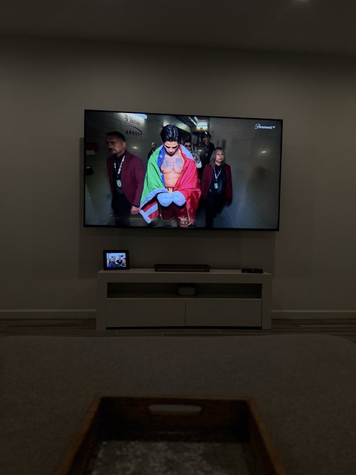 A wall-mounted TV displaying a fighter walking out draped in colorful flags, viewed from across a dark room with a TV stand below.