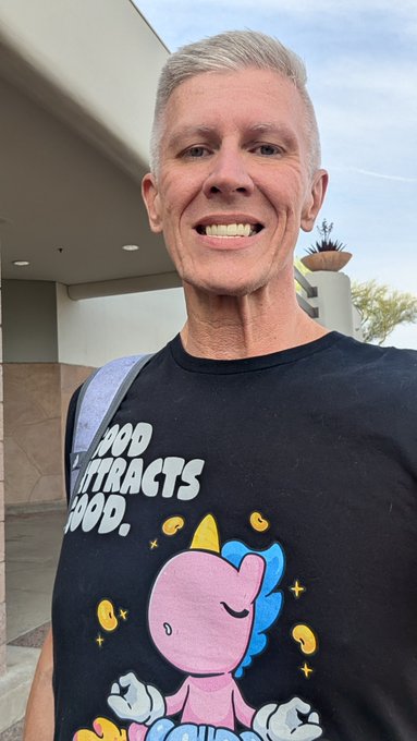 A smiling person with short gray hair wearing a black t-shirt that reads "GOOD CONTRACTS = GOOD" above a cartoon pink unicorn surrounded by coins.