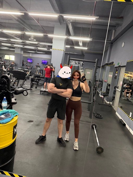 The image shows a man and woman posing together at a gym. The man has his arms crossed over his chest while wearing black shorts and a black shirt with a red logo. The woman stands next to him, also dressed in brown leggings and a black top. They are both smiling for the camera. In the background, there is another person who appears to be working out on a weight machine. A water bottle can be seen placed nearby.