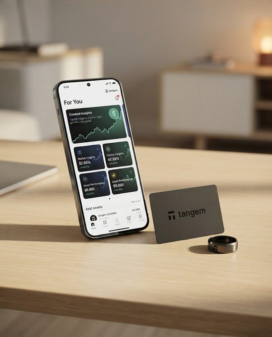 A promotional image featuring a smartphone displaying the Tangem app's 'For You' page with crypto insights and asset portfolios, sitting next to a black Tangem hardware wallet card and a ring on a wooden table.
