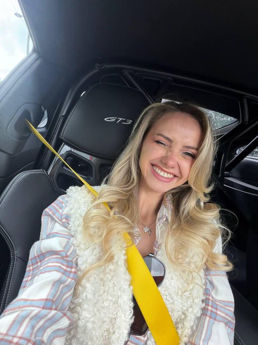 A blonde woman smiles for a selfie while sitting in the passenger seat of a Porsche GT3, identifiable by the 'GT3' logo on the headrest.