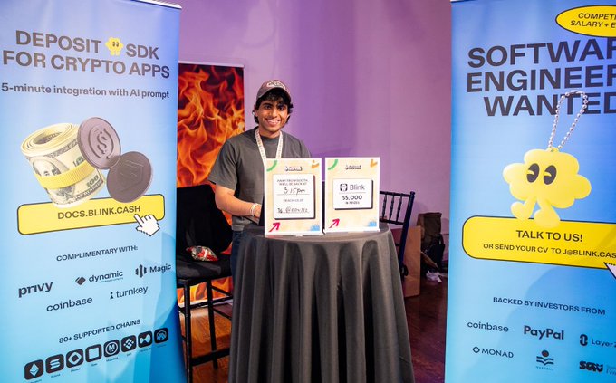 The image features a man standing behind two blue banners, one of which says "Software Engineer Wanted". The other banner has an advertisement for Bitcoin Cash XRP. In front of him are two tables with signs that say "Dogs Link" and "Talk To Us!". A chair is also visible in the scene.