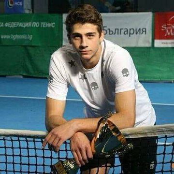 A photograph of a young male tennis player in a white shirt with skull logos, resting on a tennis net and holding a trophy, with a Bulgarian tennis federation banner in the background.