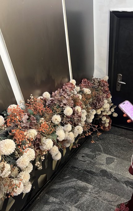 The image features a large bouquet of flowers placed against a wall with a door nearby. The flowers are arranged in various sizes and colors, creating an eye-catching display. A cell phone can be seen resting next to the flowers on the floor, suggesting that someone was using it while admiring the floral arrangement or taking a break from their activities.
