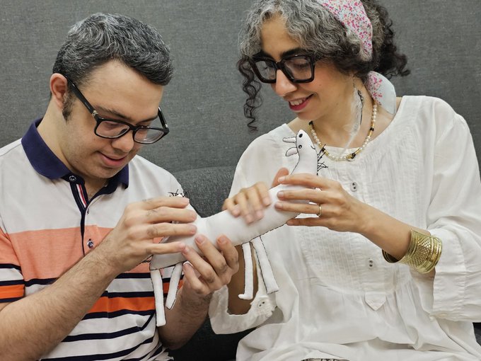 The image features a man and woman sitting together, looking at a white stuffed animal that they are holding up for each other. The stuffed animal appears to be a horse or a doll, depending on the perspective. They seem to be enjoying their time together as they interact with the toy.
