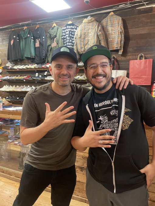 Two smiling men posing inside a sneaker and streetwear shop, with the man on the left in an olive t-shirt and the man on the right wearing a black hoodie reading 'EN HIEROGLYPH', glasses, and a green cap.