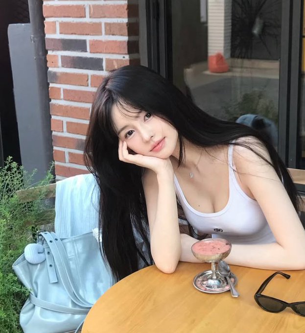 A young woman with long dark hair wearing a white tank top, posing with her chin resting on her hand at an outdoor table with a dessert cup, sunglasses, and a light blue bag beside her against a brick wall background.