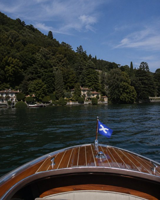 A classic wooden boat glides across a calm lake toward a hillside village, with a blue flag bearing a white cross mounted at its bow—visually confirming the "flag checked" caption as the distinctive pennant proudly flies in view.