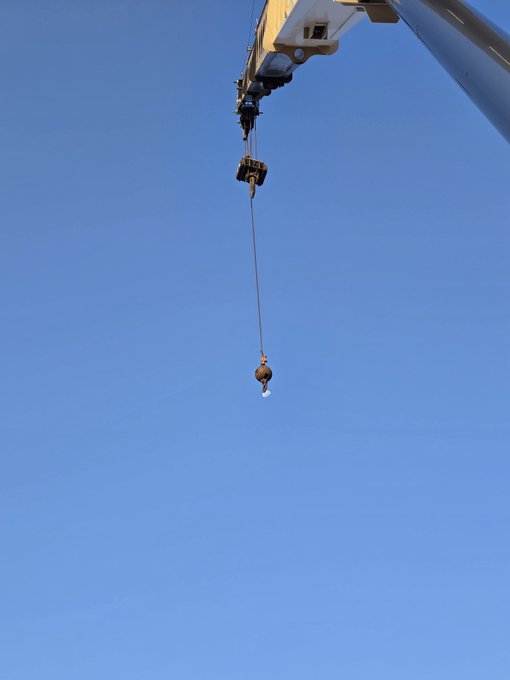 A view looking up at a construction crane's hook and cable against a clear blue sky, with what appears to be a wrecking ball or weight attached.
