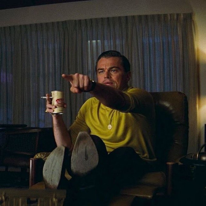 Leonardo DiCaprio, holding a paper cup and cigarette, points directly at the camera while lounging in a chair with his feet up—a still from "Once Upon a Time in Hollywood"—a visual cue to grab the viewer's attention to the article about how NFTs can yield returns beyond just deflationary token rewards.