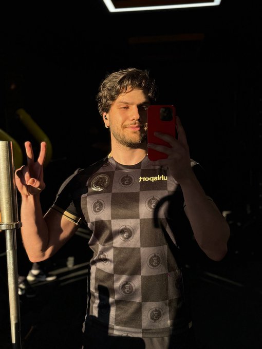 A man in a black and gray checkered sports jersey takes a mirror selfie with a red smartphone, flashing a peace sign in a dimly lit setting.