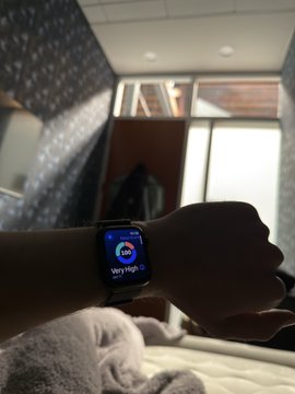 The image shows a person wearing a smartwatch with an app displaying their heart rate, which is high. The watch is placed on someone's wrist, indicating that they are actively monitoring their health or fitness data. The background features a bed, suggesting that this person might be in a bedroom setting.