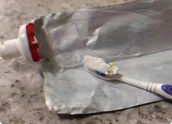 A crushed toothpaste tube lies on a counter with a spoonful of toothpaste scooped from it, humorously illustrating the POV of someone who "held" the tube too tightly and squeezed out all the contents.