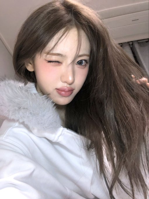 A close-up selfie of a young woman with long brown hair winking at the camera, wearing a white jacket with a fur collar.