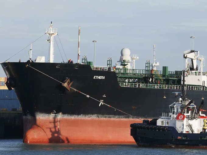 Photograph of a large black and red cargo ship named 'ETHERIA' docked at a port, accompanied by a smaller tugboat with a 'Smit' logo.