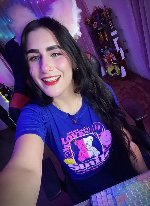 A selfie of a smiling woman with bunny ears and a blue t-shirt featuring a pink teddy bear design that says 'Spread love & kindness', sitting at a desk with a colorful RGB keyboard.