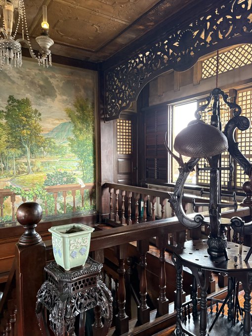 An ornate wooden interior featuring intricate carvings, a landscape mural on the wall, a decorative ceramic pot on a carved stand, and elaborate metal lanterns hanging from the ceiling.