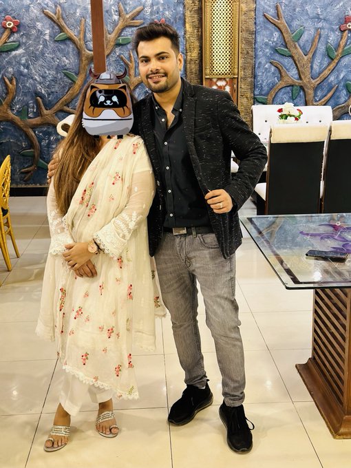 A man in a black blazer and grey jeans poses with a woman whose face is obscured by a bulldog mask; she wears a cream-colored floral embroidered dress.