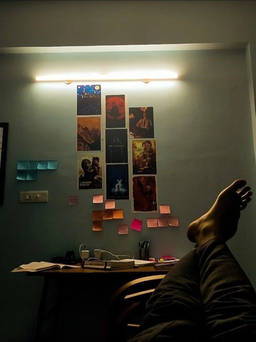 A cozy, dimly-lit room shows a person resting with their foot up, facing a wall decorated with a collage of movie and art posters above a desk scattered with sticky notes and study materials—capturing a quiet late-night work session. The intimate, studious setting reflects the "keep building" hustle of a founder burning the midnight oil on flexible compute infrastructure.