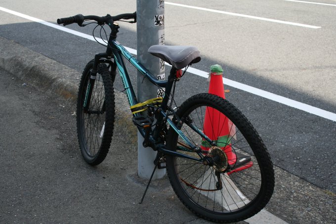 A black and teal mountain bike locked to a metal pole on a sidewalk, positioned next to a bright orange traffic cone near a street.